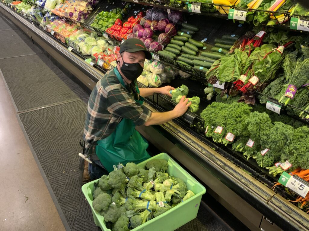 A produce clerk wearing a mask, apron, and hat looks at the camera as he loads broccoli onto the bottom shelf produce wall
