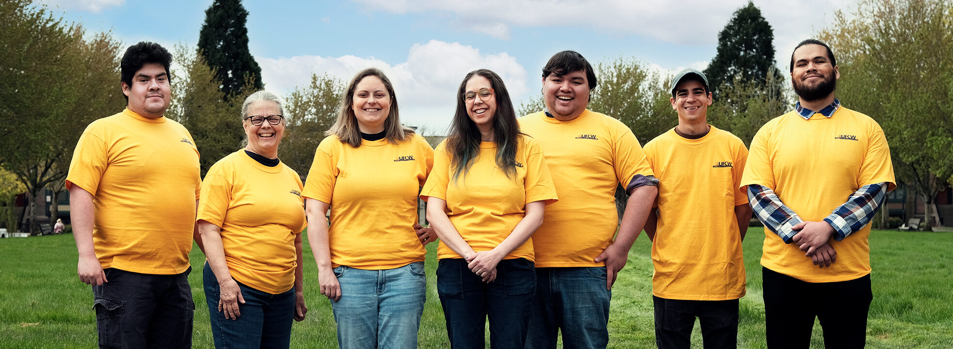 Seven members of the UFCW New Seasons team stand shoulder to shoulder outdoors, with a blue sky and fluffy clouds behind them. All are wearing UFCW gold shirts and looking at the camera proudly.